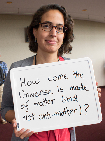 Photo of Alysia Marino holding whiteboard saying "How come the universe is made of matter (and not anti-matter)?"