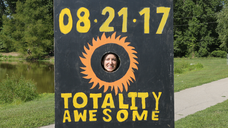 Photo of woman putting her face through wood cut out that says "08.21.17 Totality Awesome" for great eclipse
