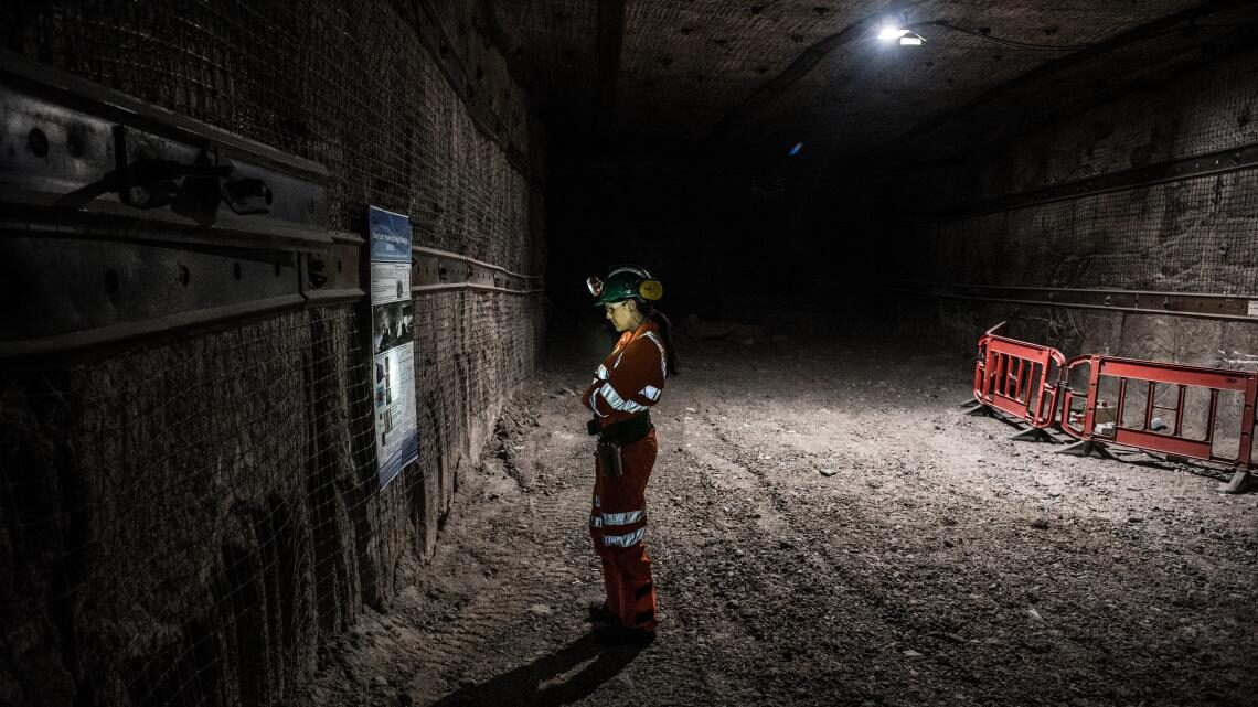 Scientist standing in a mine tunnel, reading a sign, with her face lit by a headlamp on her helmet