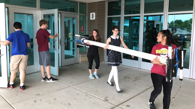 Photo of three students carrying a long detector while another holds the door