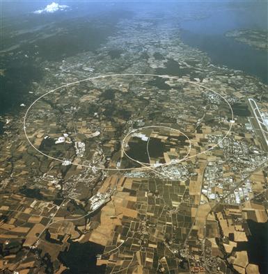 Three rings are visible, the smaller shows the underground position of the PS, the middle ring is the SPS with a circumference of 7 km and the largest ring (27 km) is that of the former LEP accelerator with part of Lake Geneva in the background. Courtesy of Cern.