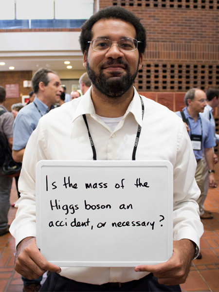 Photo of Peter Onyisi holding a whiteboard that says "is the mass of the Higgs boson an accident, or necessary?"