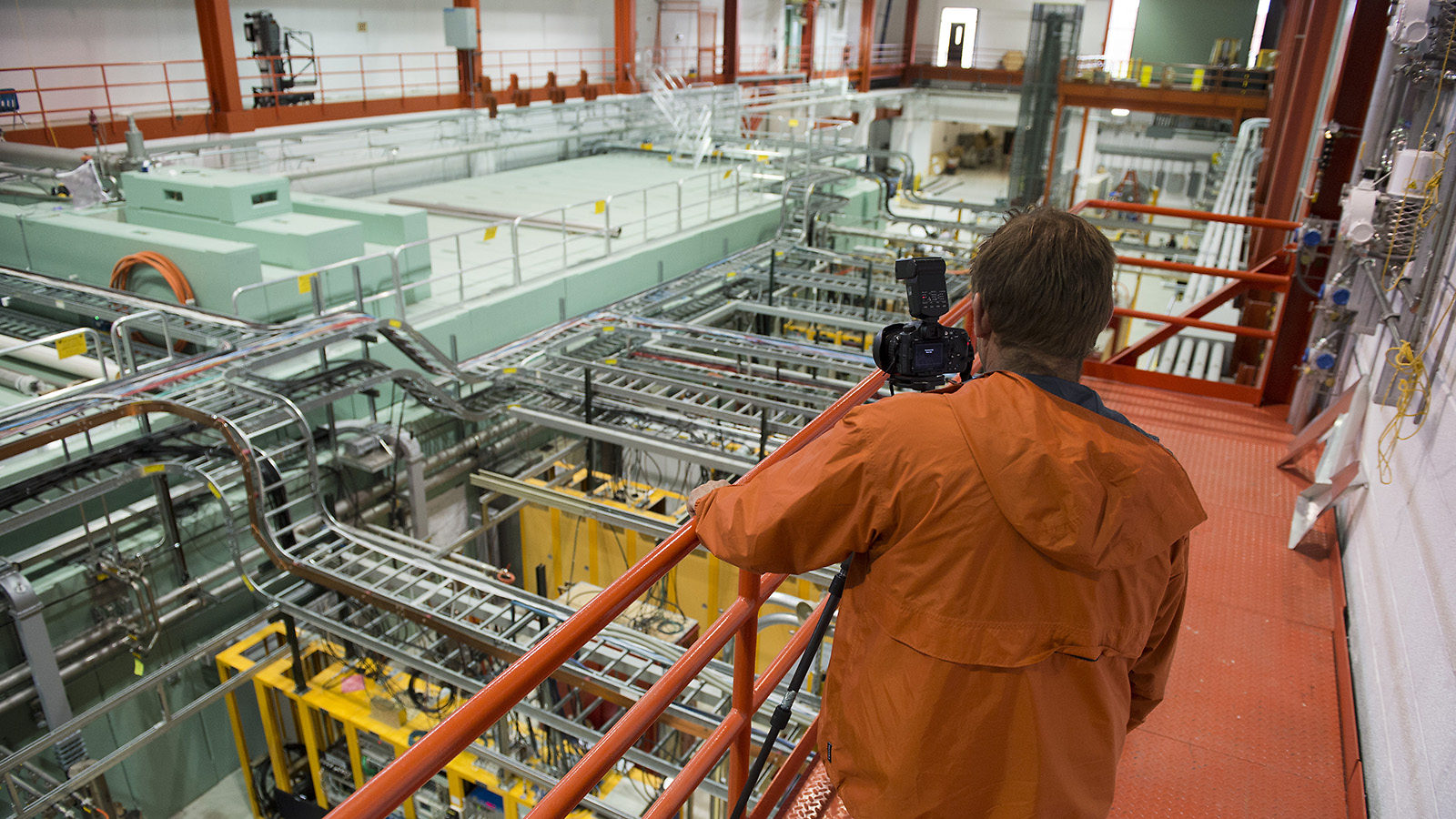 Photo of person wearing an orange jacket takes a picture of a room full of equipment from a catwalk.