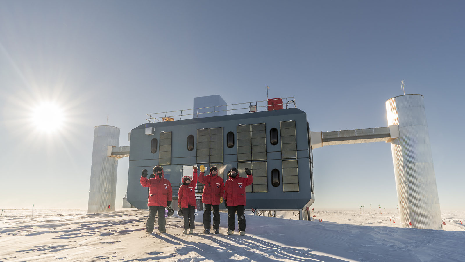 Photo of four researchers in red coats standing on white snow at the South Pole in front of a building