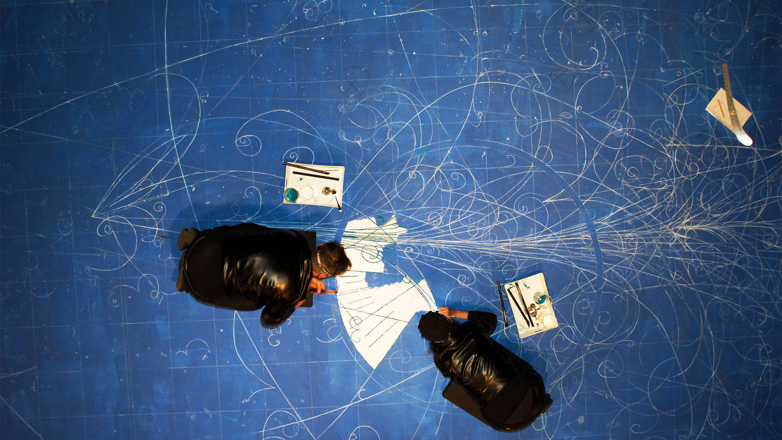 Overhead view of artists kneeling on a blue surface and creating an image of bubble chamber tracks with white sand