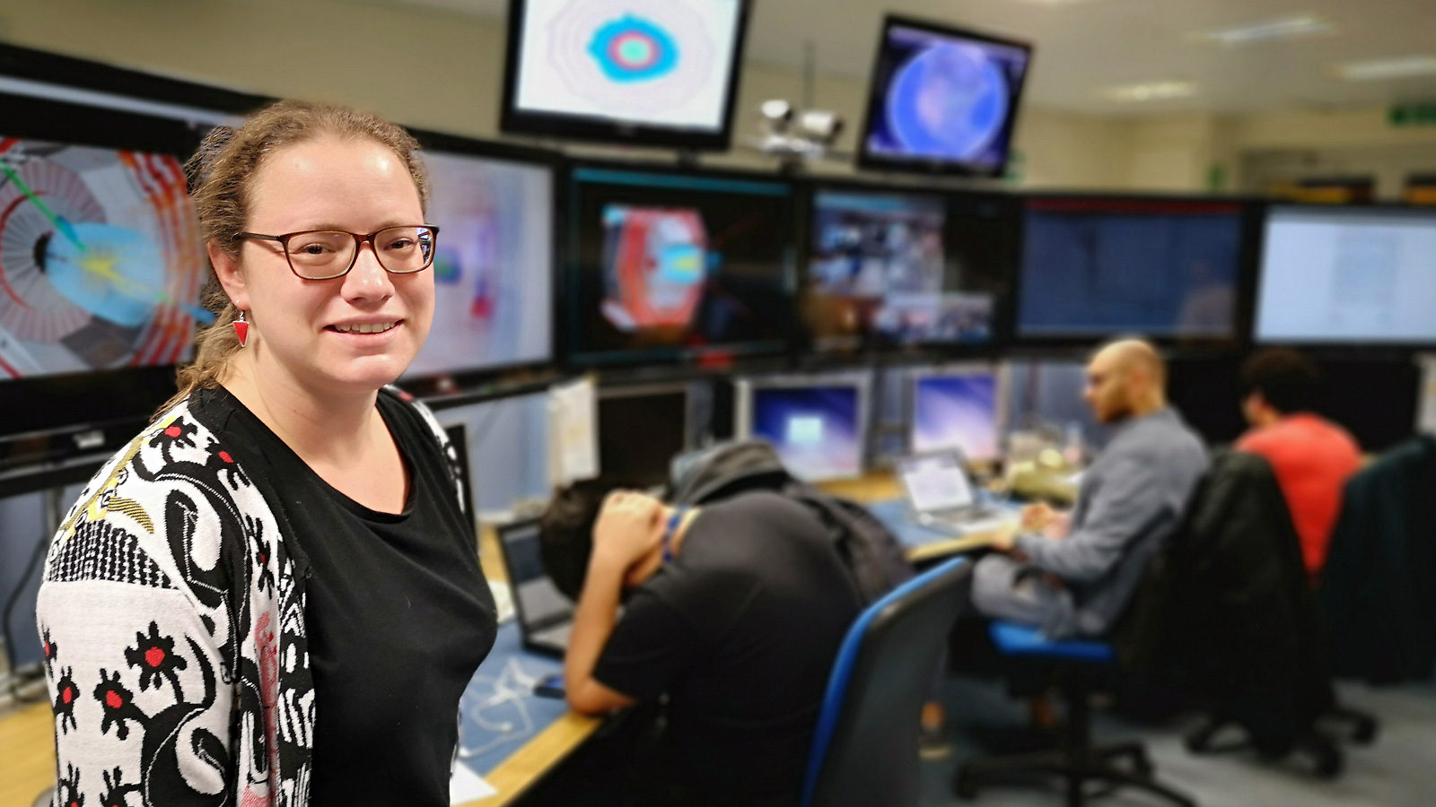 Image of a woman in glasses standing in front of several screens in a control room