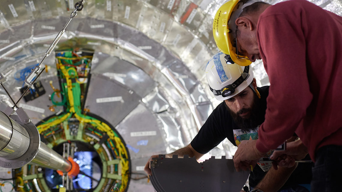 Two scientists in hard hats work with a mock-up next to the CMS detector.