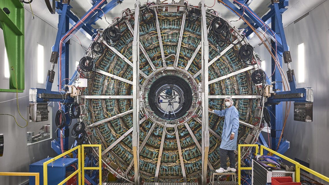 Image of a person in clean-room gear standing in front of a detector component that is twice as tall