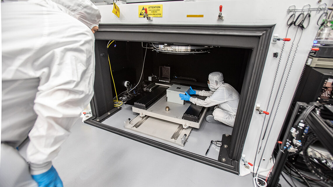 Photograph of two scientists in white clean room outfits placing a box under the LSST Camera focal plane