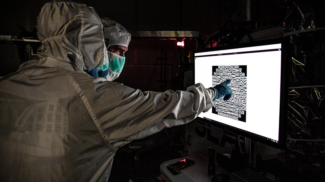 Photograph of two scientists in clean room garb looking at an image on a screen
