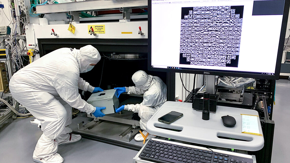Photograph of two scientists in clean room garb moving the box from under the LSST focal plane