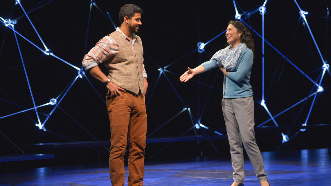 A scene from Constellations: a man and woman on stage she is reaching her hand out to him for a handshake