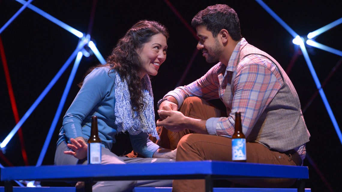 A scene from Constellations: a man and woman on stage both are sitting on a bench with beer