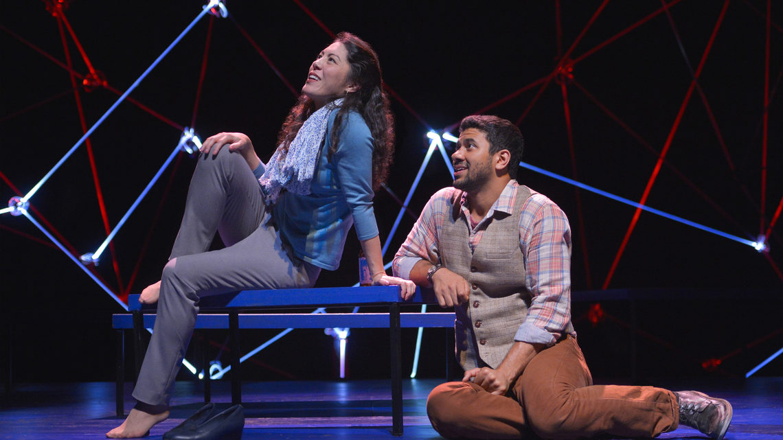 A scene from Constellations: a man and woman are on stage, she is sitting on a bench he is sitting on stage looking at her