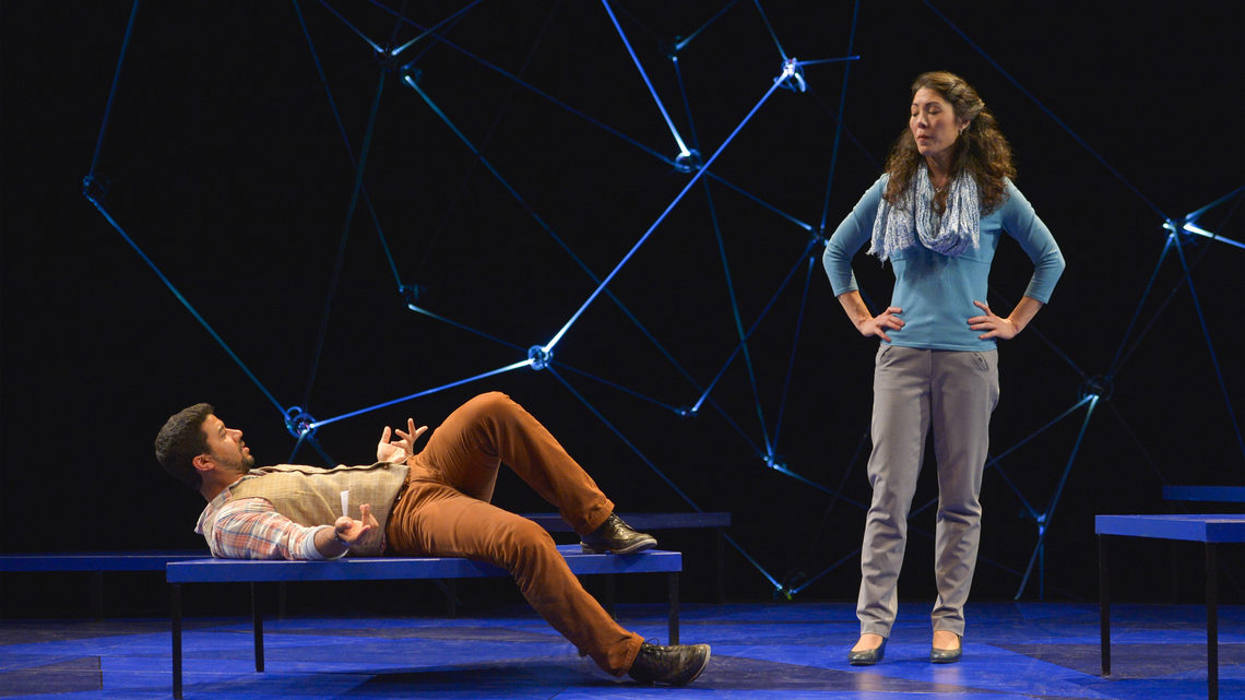 A scene from Constellations: a man and woman on stage he is laying down on a bench, she is standing with hands on her hips