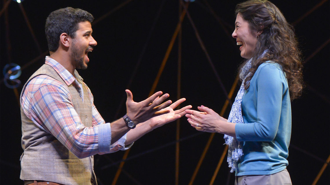 A scene from Constellations: a man and woman on stage he explaining something to her, she is laughing 