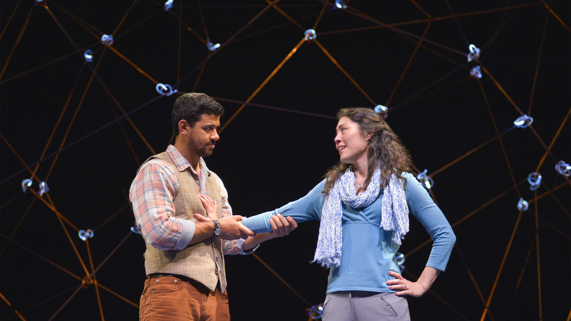 A scene from Constellations: a man and woman on stage. He is holding her arm against his chest