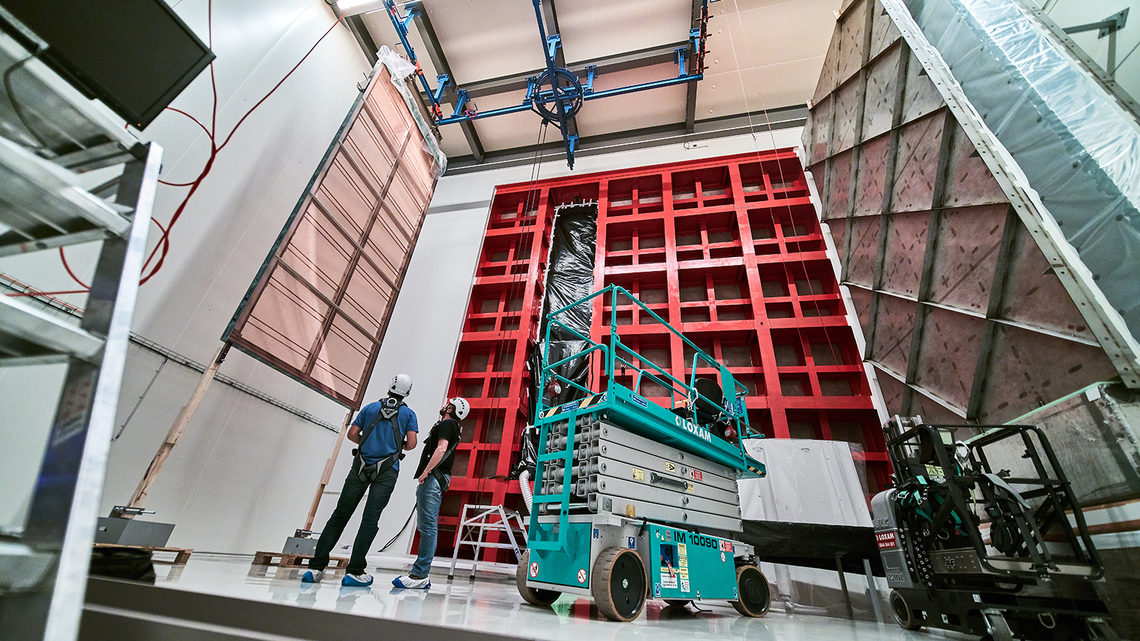 Technicians look up at an APA hanging near the red ProtoDUNE frame
