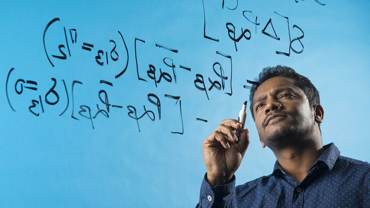 Photo of Jogesh Rout writing equations on a clear panel, blue background