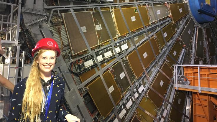 Photograph of Jannicke Pearkes, a blonde woman in a CERN hard hat, standing next to the ATLAS detector