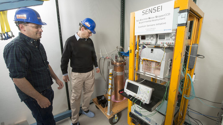 Two scientists in hard hats stand next to a cart holding detector components.