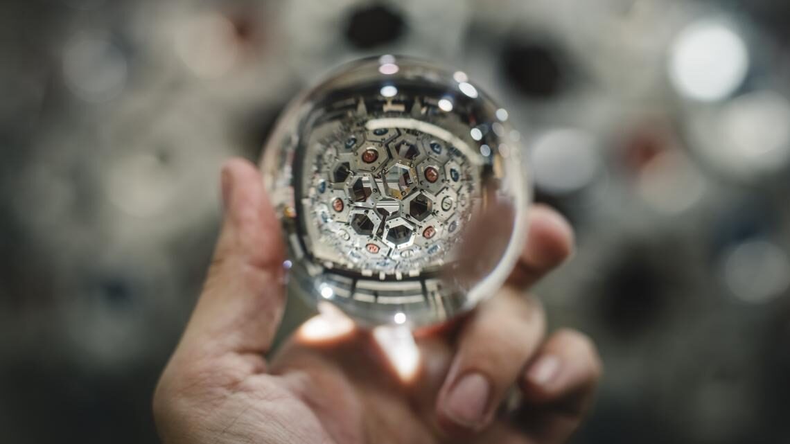 Photo of a hand holding a glass ball, through which one can see a silver particle detector 